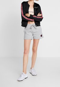 Svart bomberjacka med röda och vita randiga detaljer, ljusbeige crop top, gråa sweatshorts med logotyp och vita sportskor.