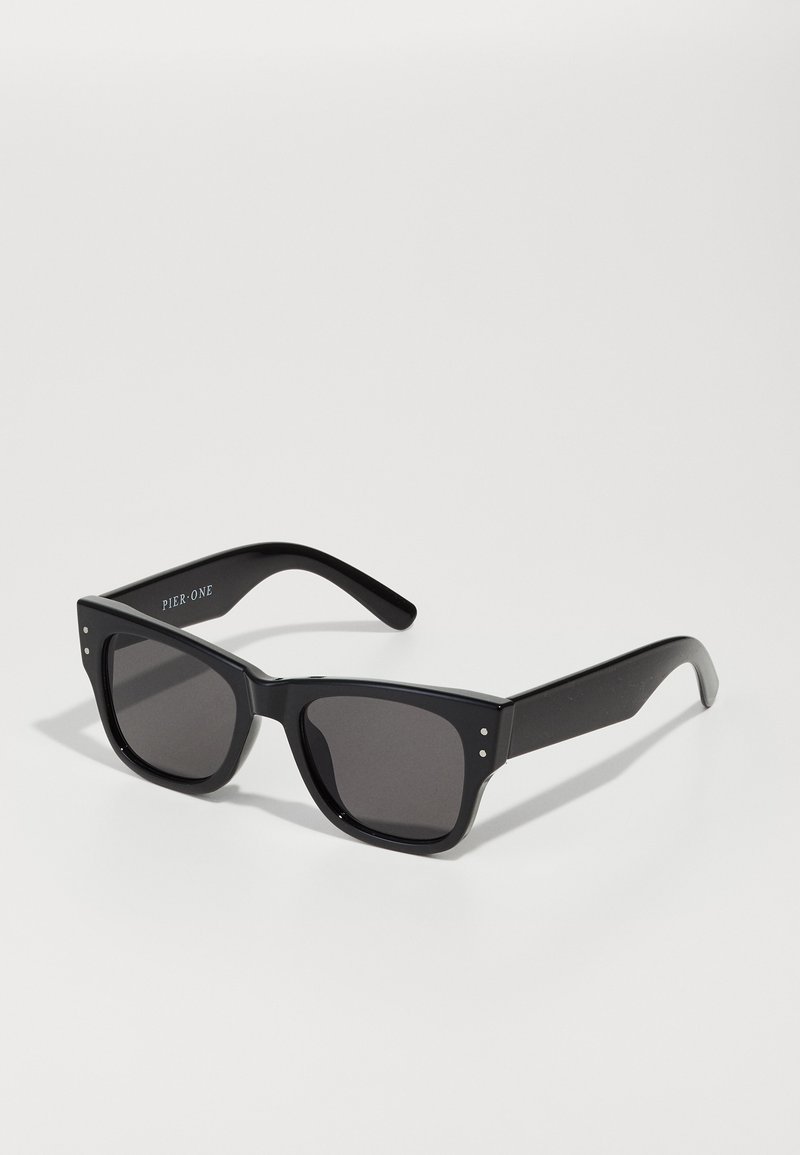 Ochelari de soare pătrați negri cu lentile fumurii, cadru gros din plastic și patru dibluri metalice pe față. Brațele sunt complet negre și elegante.