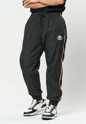 Person iført sorte Kappa træningsbukser med orange sidelommer og sorte og hvide sneakers, stående med armene over kors.