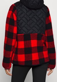 Czerwona i czarna polarowa kurtka w kratę z pikowanym czarnym panelem na górnej części pleców i kapturze. Posiada długie rękawy i luźny krój.