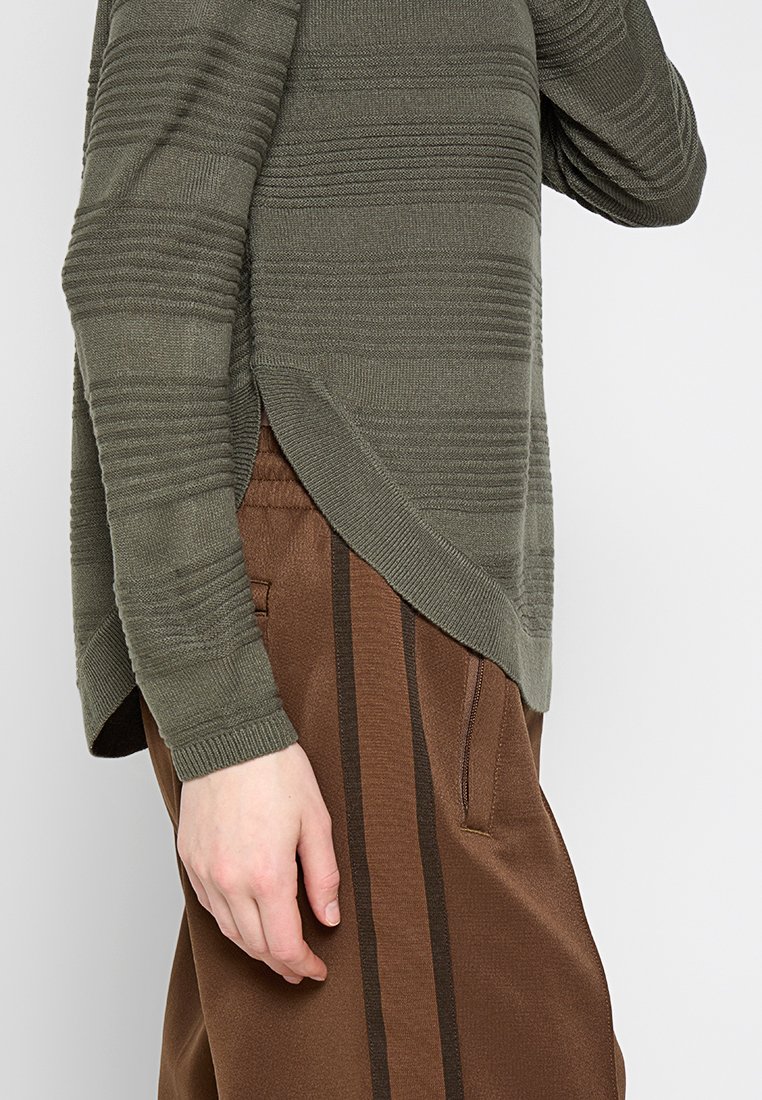 Vista laterale di una persona che indossa un maglione verde oliva testurizzato con orlo asimmetrico e pantaloni marroni con strisce verticali scure.