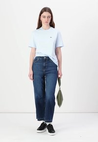 Tricou albastru deschis cu mânecă scurtă, cu sigla Lacoste, asortat cu blugi evazați închiși la culoare cu talie înaltă și adidași negri, ținând o geantă verde.