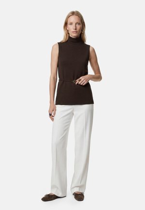 Kvinde iført sort ærmeløs rullekravetop med tyndt bælte, hvide vidde bukser, brune flade sko og guld creoløreringe mod hvid baggrund.