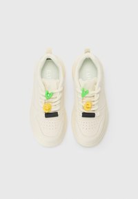 Cremfarbene Sneakers mit strukturierten Oberflächen, durchlöchtem Zehenbereich und einzigartigen Schnürsenkeln, die mit grünen und gelben dekorativen Charms versehen sind.