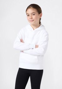 Chica con una sudadera blanca de Champion, con los brazos cruzados, sonriendo levemente, de pie contra un fondo claro y liso.
