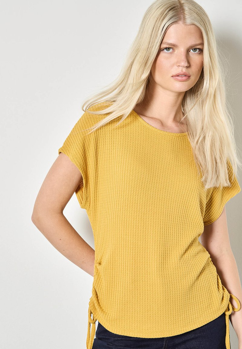 Haut jaune à manches courtes avec texture gaufrée, muni de cordons de serrage sur les côtés, offrant une coupe décontractée et un col rond.