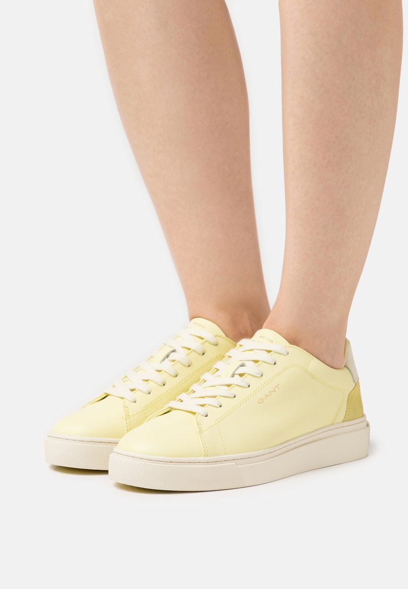 Sneakers in pelle gialla con lacci bianchi, texture liscia, punta tonda e dettaglio del logo. Presenta una suola di colore chiaro e un dettaglio del tallone a contrasto.