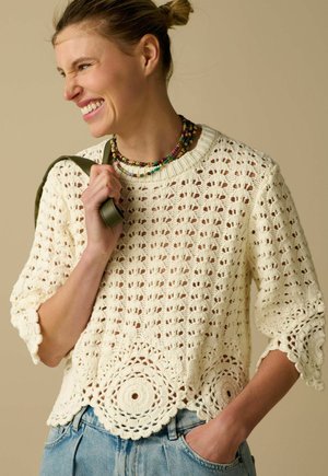 Femme souriante, portant un pull crochet crème avec des motifs circulaires, des colliers de perles superposés, un jean bleu clair et tenant une bandoulière vert olive.