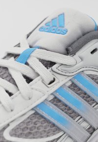 Grå sportskor med vita och blå detaljer, med en meshtopp, texturerat material och tre blå ränder på sidan.