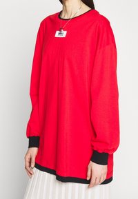 Kvinna som bär en oversized ljusröd långärmad tröja med svarta manschetter och nederkant över en ljus plisserad kjol, med ett hängande halsband.