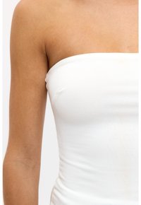Close-up van het bovenlichaam en de arm van een persoon in een witte strapless top tegen een effen achtergrond.
