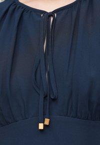Μπλούζα σε ναυτικό μπλε με πλισέ ύφασμα, που διαθέτει δέσιμο με θηλιά στο ντεκολτέ και τετράγωνες χρυσές λεπτομέρειες στα άκρα των δεσιμάτων.