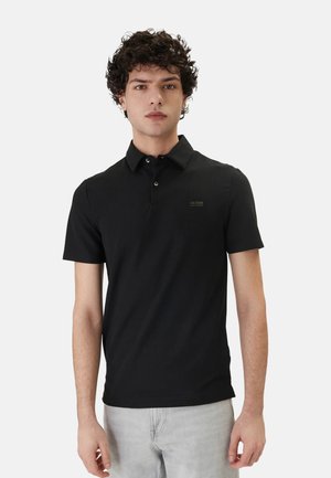 Giovane uomo con capelli ricci che indossa una polo nera aderente a maniche corte e pantaloni grigio chiaro, in piedi davanti a uno sfondo bianco.