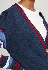 Cardigan blu navy con accenti a strisce rosse e blu, chiusura con bottoni e tessuto a maglia testurizzata. Presenta una vestibilità comoda e un design aperto nella parte anteriore.