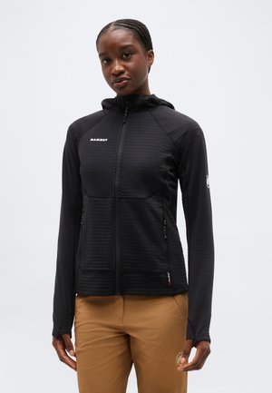 Mladá žena v čiernej mikine na zips s kapucňou a v béžových nohaviciach stojí pred jednoliatym svetlým pozadím.