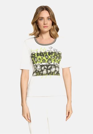 Vrouw met halflang blond haar, gekleed in een wit shirt met groene en grijze luipaardprint en de tekst "keep life simple" op de voorkant.