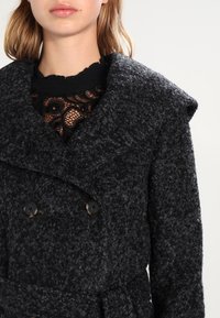 Abrigo negro de doble botonadura con una tela texturizada similar a la lana. Presenta un amplio collar y cinturón, con botones oscuros y una blusa de encaje en capas debajo.