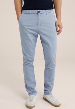 Mężczyzna noszący jasnoniebieskie spodnie slim-fit z zapięciem na guzik i zamek, w połączeniu z białymi trampkami i ciemnym topem.