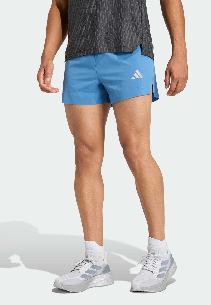 Modré športové šortky s hladkým povrchom, s bočnými rozparkami a bielym logom Adidas. Model nosí sivé a biele bežecké topánky.