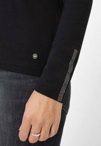 Hand, die einen silbernen Ring trägt, liegt an der Seite, bekleidet mit einem schwarzen langärmligen Oberteil mit silbernem Nietenstreifen am Ärmel und einem kleinen runden Metalllogo in der Nähe des Saums.