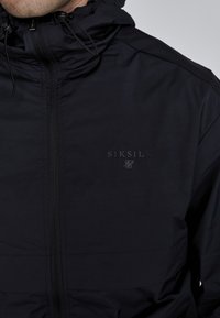 Black waterproof jacket with a high collar, front zipper, cinch drawstrings, and a subtle logo embossed on the chest. Smooth texture.