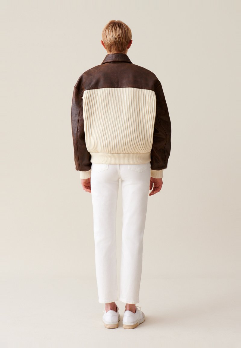 Braune Lederjacke mit cremefarbenem geripptem Stoff und hohem Kragen, kombiniert mit weißen Slim-Fit-Hosen und weißen Sneakers. Einfaches Design, keine Muster.