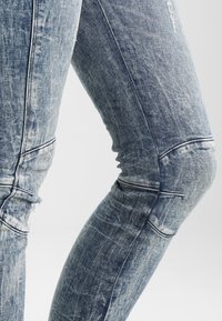 Vaqueros de denim azul con un diseño desgastado, forma ajustada y costuras de rodilla visibles. La tela presenta diferentes tonalidades y texturas.