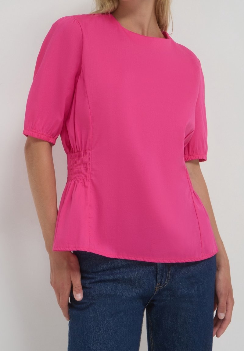 Femme portant un chemisier rose vif à manches courtes, avec une taille froncée, et un jean bleu, debout devant un fond uni clair.