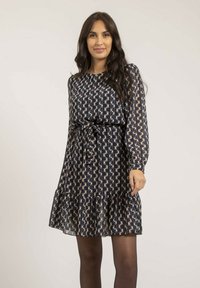 Robe bleu marine à manches longues, avec une ceinture à nouer à la taille et un ourlet à volants. Présente un motif géométrique abstrait en beige et crème.