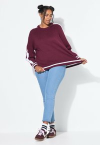 Maglione a maglia bordeaux con righe bianche, abbinato a jeans skinny azzurri chiari con dettagli borchiati. Il modello indossa scarpe scure e calzini a righe.