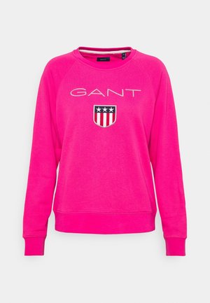 Sweat-shirt rose vif à manches longues avec logo GANT et un emblème en forme de bouclier comportant trois étoiles ainsi que des rayures rouges et blanches sur la poitrine.