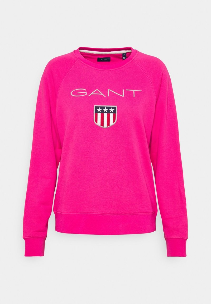 Sweat-shirt rose vif à manches longues avec logo GANT et un emblème en forme de bouclier comportant trois étoiles ainsi que des rayures rouges et blanches sur la poitrine.