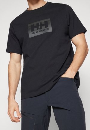 Mand iført en sort Helly Hansen t-shirt med logo på brystet og mørke bukser, stående med den ene hånd i lommen mod en ensfarvet baggrund.