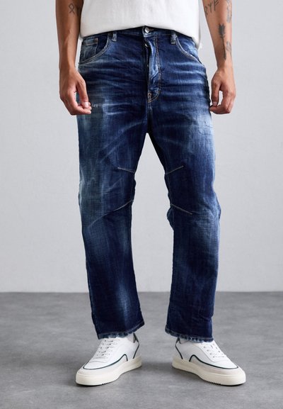 Donkerblauwe jeans met een versleten uitstraling, recht gesneden, vijf zakken en gele stiksels, gecombineerd met witte sneakers.