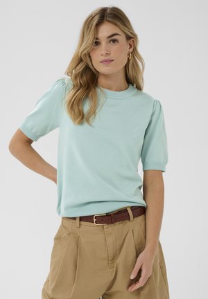 Maglione verde menta chiaro a maniche corte con scollo tondo e dettaglio a spalla gonfia, abbinato a pantaloni beige chiaro a vita alta e cintura marrone.