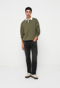 Olivgrüner Pullover mit weißem Kragen, dunkle Jeans und beige Sneakers. Leicht strukturiertes Material, lässige Passform, steht vor einem schlichten Hintergrund.