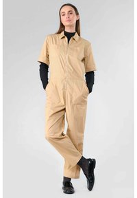 Jumpsuit beige à manches courtes avec une fermeture éclair devant, deux poches poitrine et une coupe décontractée. Associé à un haut noir à manches longues et des chaussures noires brillantes.