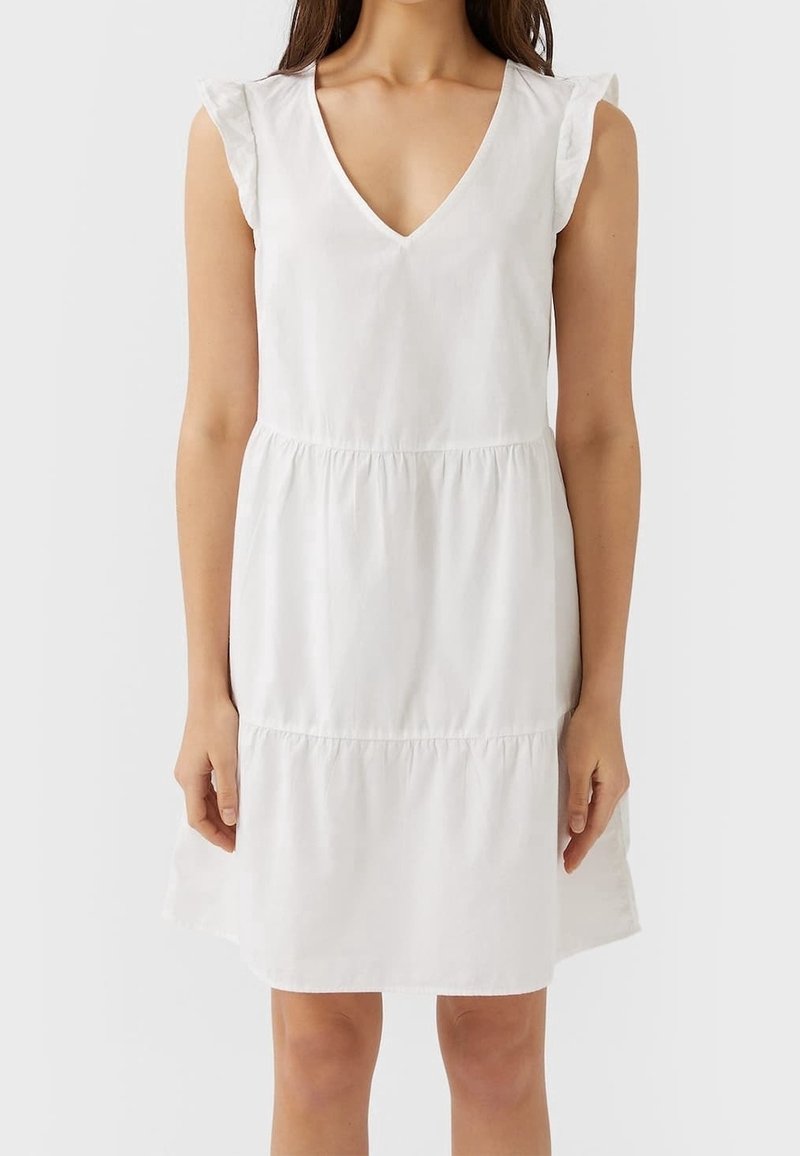 Femme portant une robe blanche sans manches avec un décolleté en V et des épaules à volants, debout devant un fond clair uni.