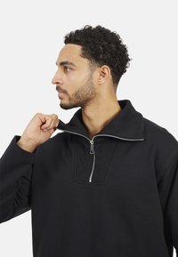 Pull noir avec un col haut et une fermeture éclair quart. Présente des détails côtelés au niveau du col, des manches longues et une texture douce.