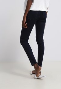 Una persona está de pie vistiendo jeans ajustados oscuros y sandalias plateadas, con la espalda hacia el espectador, contra un fondo sencillo.