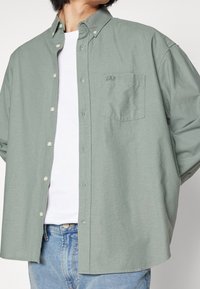 Chemise boutonnée vert clair en tissu doux, avec une poche poitrine ornée du logo GAP, et une coupe décontractée. Portée par-dessus un t-shirt blanc.