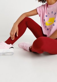 Červené legíny s hladkou texturou, spárované s růžovým tričkem s grafikou a světle růžovými teniskami s černými akcenty.