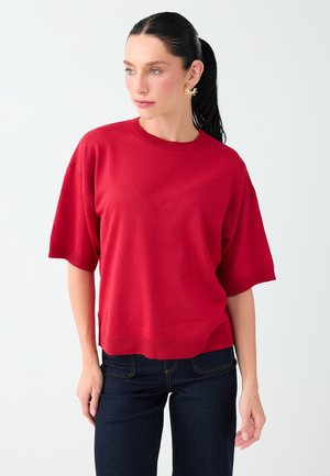 Femme avec une queue de cheval noire portant un pull rouge à manches courtes, un jean foncé et des boucles d'oreilles dorées en forme de soleil, regardant sur le côté.