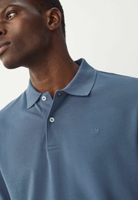 Polo marškiniai tamsiai mėlynos spalvos, pagaminti iš tekstūruoto audinio. Turi klasikinį apykaklę, du mygtukus ir mažą siuvinėtą logotipą ant krūtinės.
