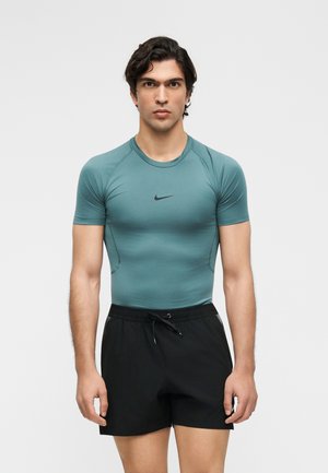 Jeune homme portant un t-shirt Nike ajusté turquoise à manches courtes et un short de sport noir avec cordon, debout devant un fond clair uni.