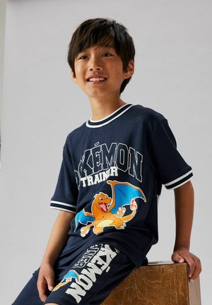 Lächelnder Junge sitzt auf einer Holzkiste und trägt ein marineblaues Pokémon-Trainer-Outfit mit Charizard-Grafik auf Shirt und Shorts.