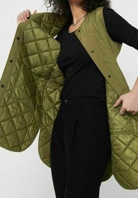 Oliwkowa pikowana kamizelka z zapięciem na guziki, z wewnętrzną miękką teksturą i lekkim, gładkim zewnętrzem, noszona na czarnej bluzce.