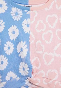 Színes felső, amelynek egyik oldala kék, fehér margarétákkal, a másik oldala pedig rózsaszín, szívmintákkal díszítve. Puha anyag, bordás nyakkivágással és varrásokkal.