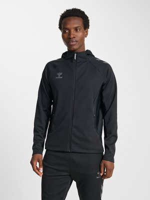 Mann trägt eine schwarze Kapuzenjacke mit Reißverschluss und passende schwarze Hose, steht entspannt mit den Händen an den Seiten vor einem einfarbigen Hintergrund.