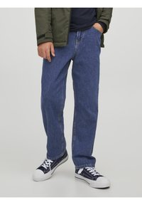 Persona che indossa jeans blu a gamba dritta, sneakers nere con lacci bianchi, una giacca verde scuro e una maglietta nera, in piedi con una mano in tasca.